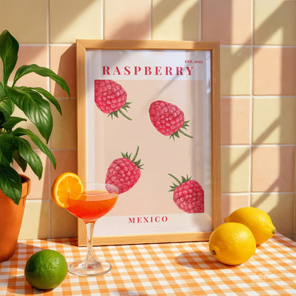 quadro emoldurado com ilustrações de framboesas em fundo claro, com moldura de madeira clara, acompanhado de copo de drink e frutas cítricas sobre mesa xadrez