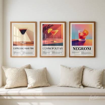 composição de três quadros decorativos com coquetéis coloridos em moldura de madeira clara, incluindo espresso martini, cosmopolitan e negroni, dispostos sobre uma parede clara ao lado de almofadas bege