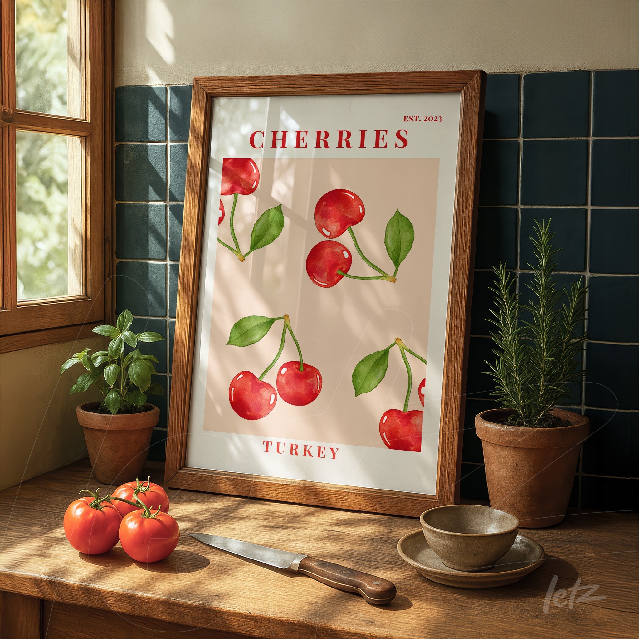 quadro em moldura de madeira clara com ilustração de cerejas sobre fundo rosa, exibido em uma mesa de madeira com tomates vermelhos e uma faca ao lado