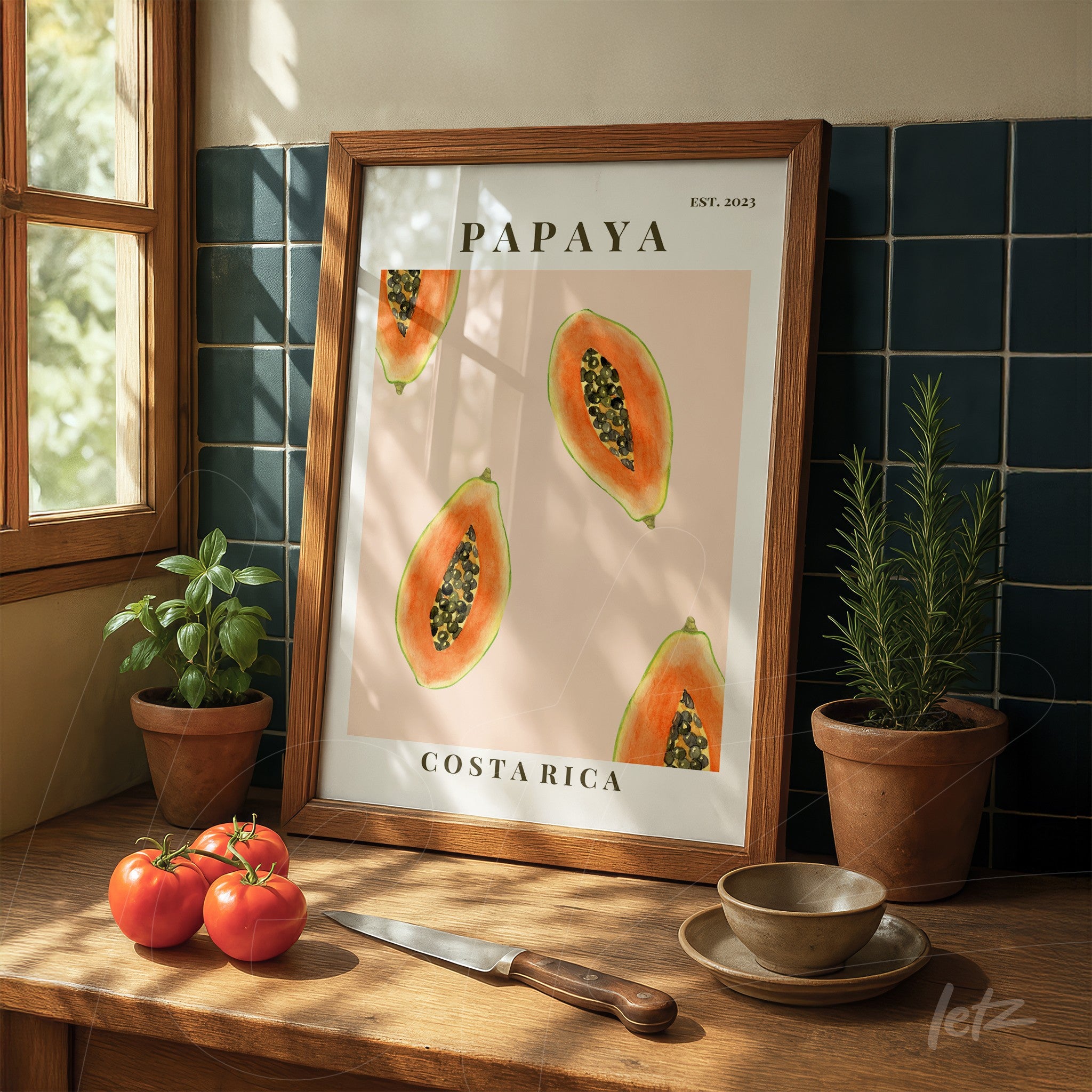 quadro em moldura de madeira clara com ilustração de papaia em fundo rosa, exposto em mesa de madeira com ervas e tomates