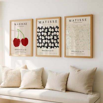 composição de três quadros em moldura de madeira clara com ilustrações de flores e frutas sobre parede branca, apoiados acima de um sofá com almofadas
