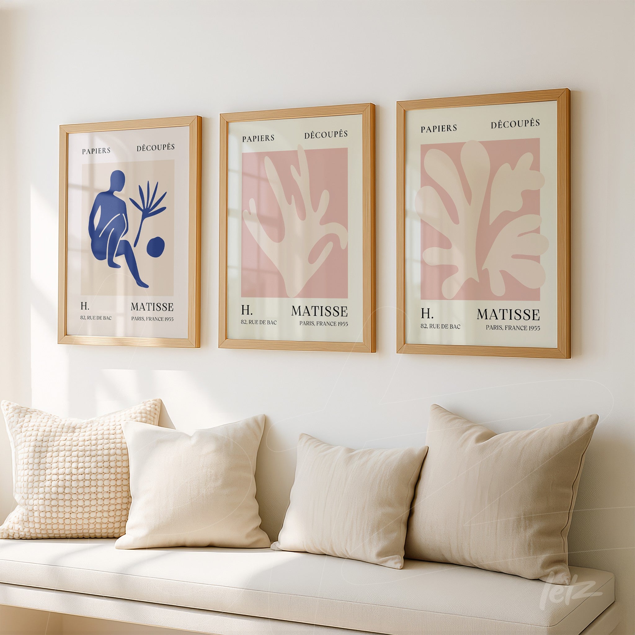 três quadros decorativos com recortes de papel em moldura de madeira clara, representando formas abstratas de plantas em tons de azul e rosa, exibidos sobre uma parede clara acima de almofadas beige