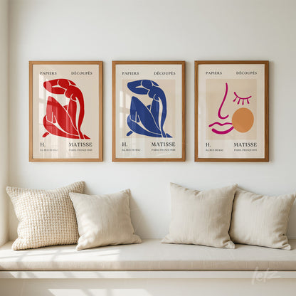 trio de quadros com arte em estilo minimalista, molduras de madeira clara, representando figuras humanas em cores vibrantes, dispostos em uma parede bege sobre um sofá com almofadas claras