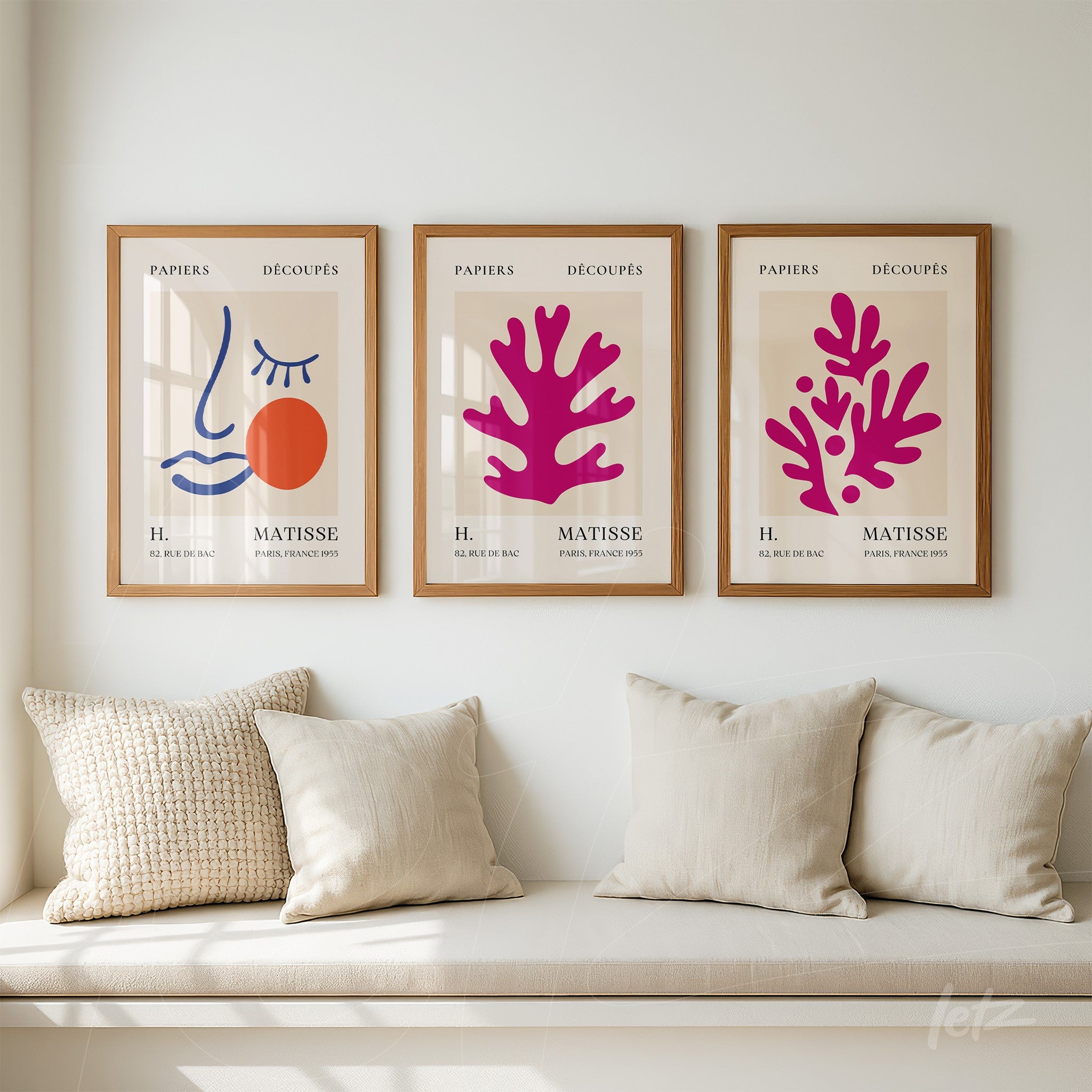 composição com três quadros decorativos abstratos em moldura de madeira clara, representando um rosto minimalista, formas orgânicas em rosa e uma composição gráfica em azul e laranja, expostos sobre parede clara