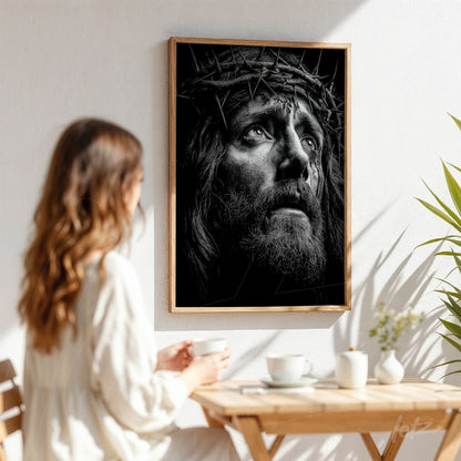 quadro em preto e branco de retrato de Jesus com coroa de espinhos em moldura de madeira clara, exposto em parede branca com planta ao lado