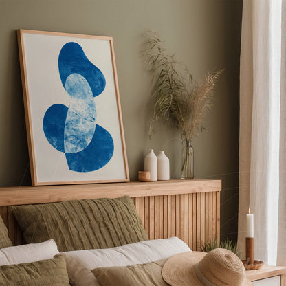 quadro com arte abstrata em formas azuis sobre fundo claro, moldura de madeira clara, exibido em ambiente minimalista com parede verde e elementos naturais como vasos e plantas