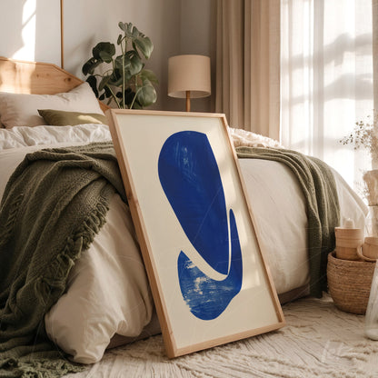 quadro moderno com forma abstrata em azul em moldura de madeira clara, apoiado na cama com lençóis brancos e cobertor verde
