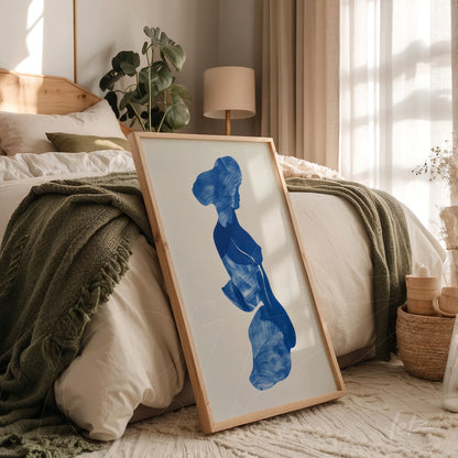 quadro de arte abstrata em azul com figura feminina em moldura de madeira clara, apoiado em cama com roupa de cama neutra