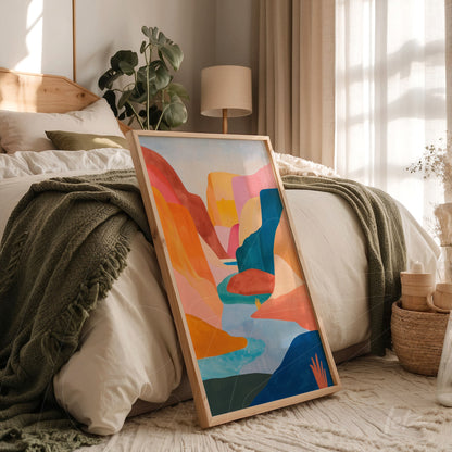 quadro com arte abstrata de paisagens em moldura de madeira clara apoiado ao lado de uma cama em um quarto iluminado