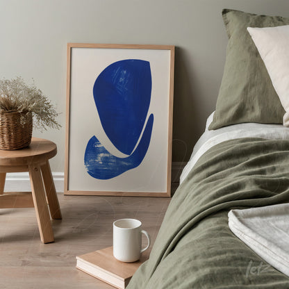 quadro emoldurado com arte abstrata em azul, apoiado ao lado de uma cama com roupa de cama em tons verde e beige