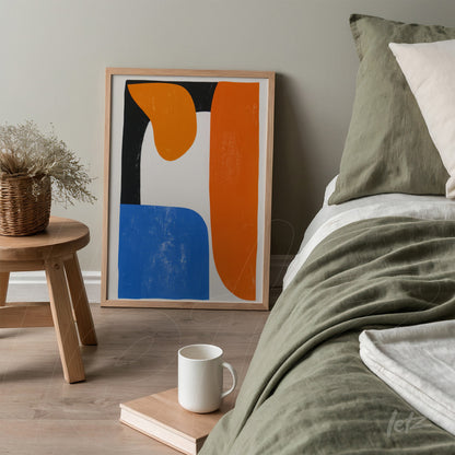 quadro com arte abstrata nas cores laranja, azul e branco em moldura de madeira clara, apoiado em mesa ao lado de uma cama com roupa de cama em tons neutros