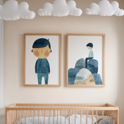 composição de quadros com ilustrações infantis de menino em azul e criança sentada sobre pedras, moldura em madeira clara, exibidos em ambiente de quarto de bebê com nuvens decorativas no teto