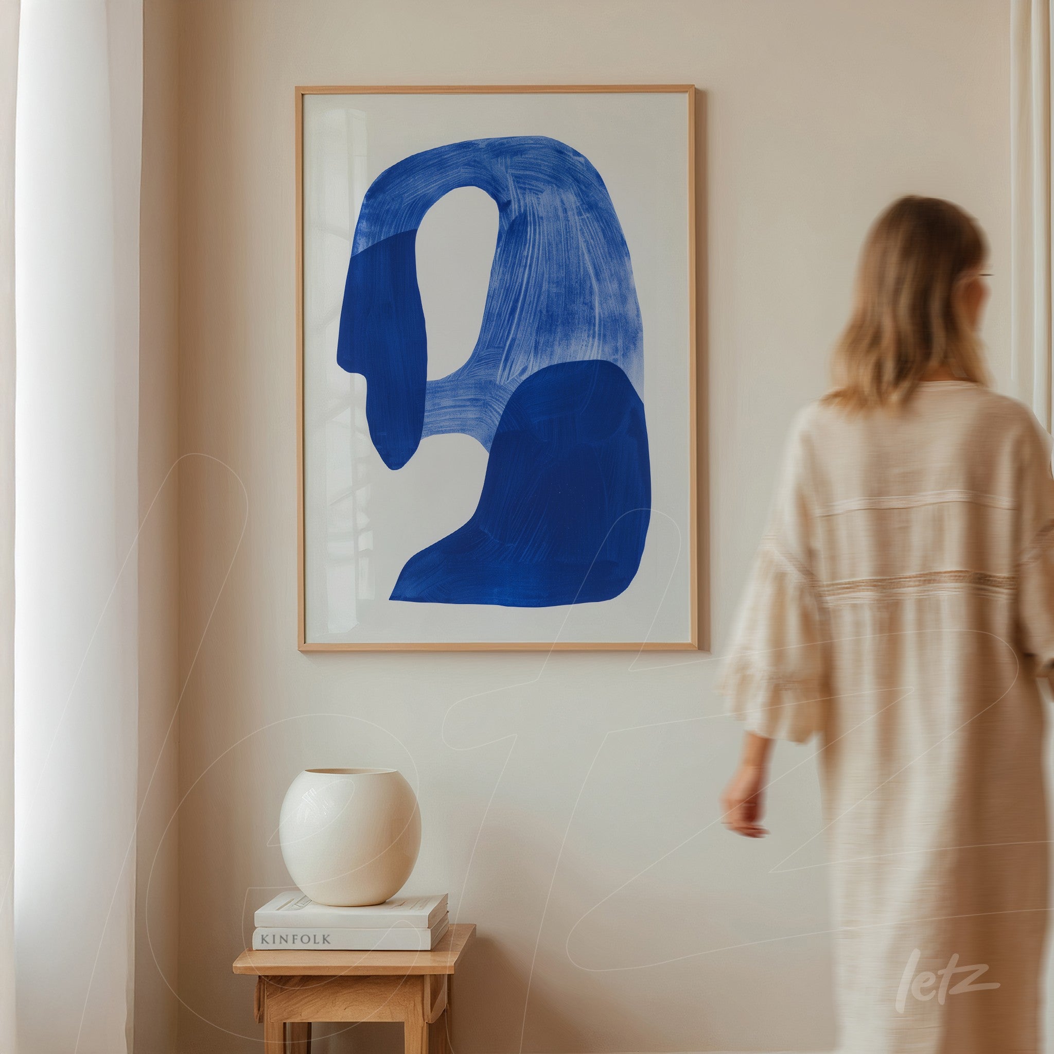 quadro abstrato em moldura clara com formas azuis em fundo branco, exibido em parede clara ao lado de um vaso