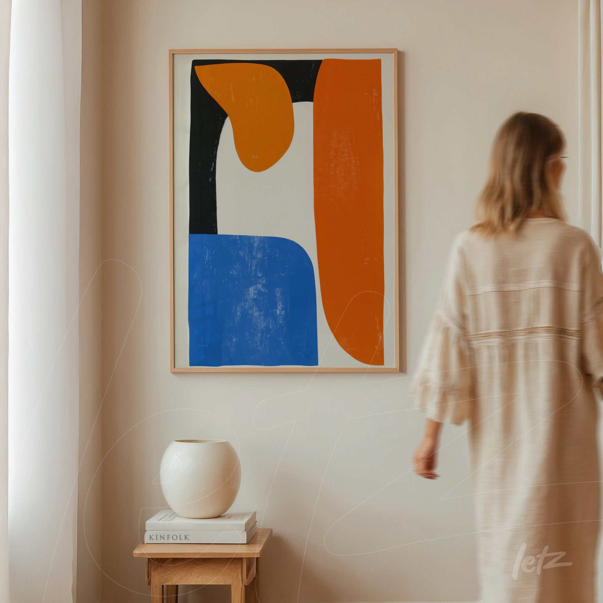 quadro abstrato em moldura clara com formas geométricas nas cores laranja, azul e preto, exibido em parede neutra ao lado de vaso branco