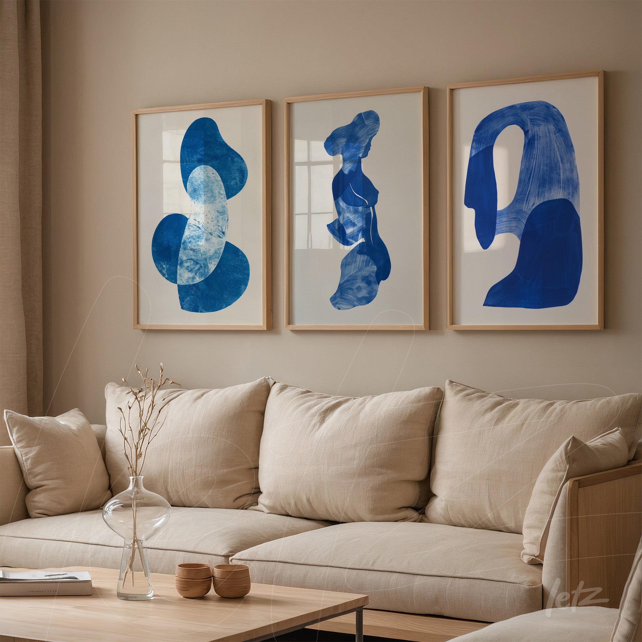 composição de três quadros com formas abstratas em tons de azul em moldura clara, exibidos sobre parede neutra ao lado de sofá acolhedor