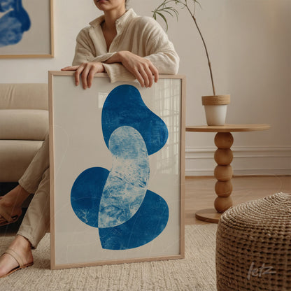 quadro com arte abstrata em tons de azul em moldura clara, apoiado sobre mesa de madeira ao lado de mulher vestindo suéter