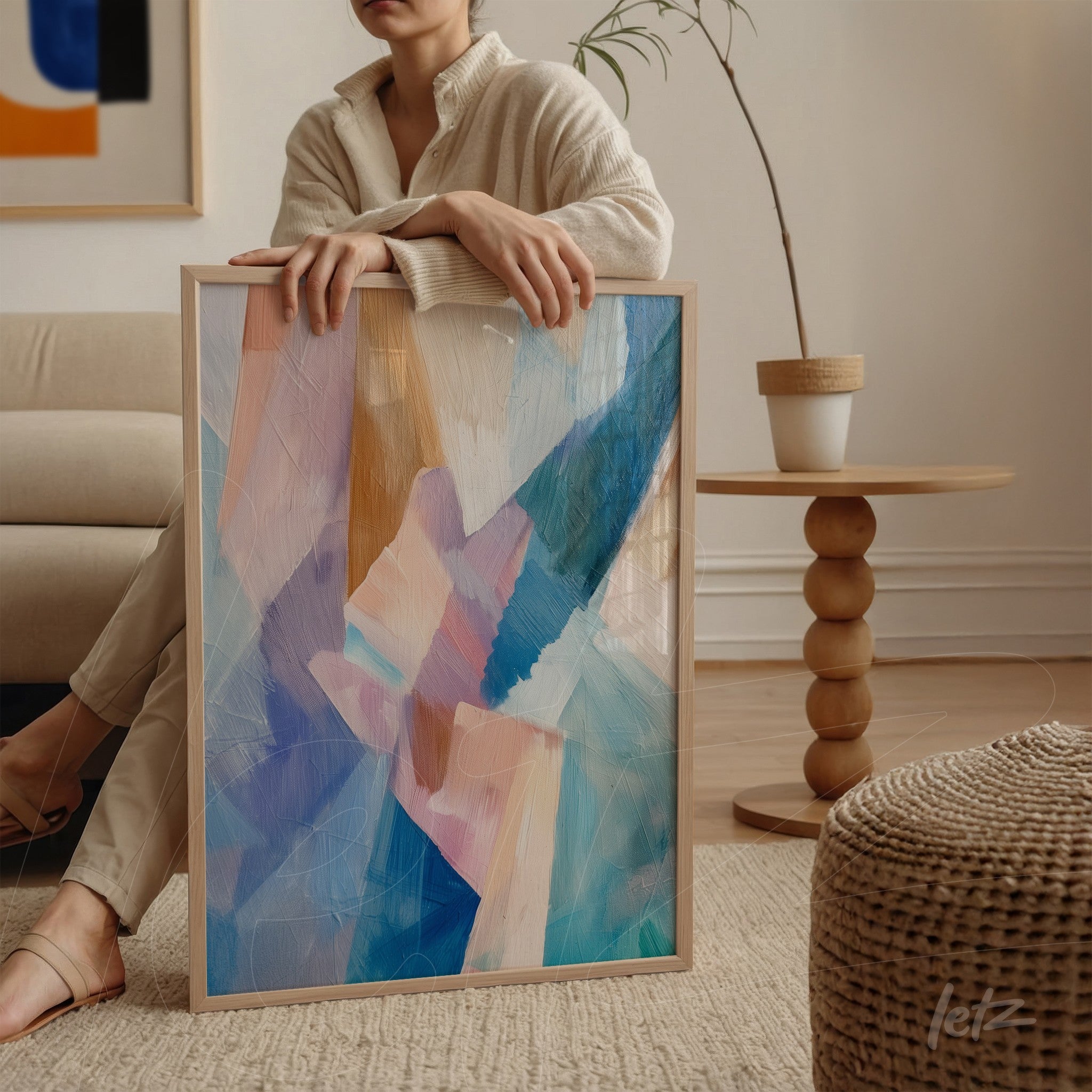 quadro de arte abstrata em tons de azul e rosa com moldura clara, apoiado em mesa de madeira
