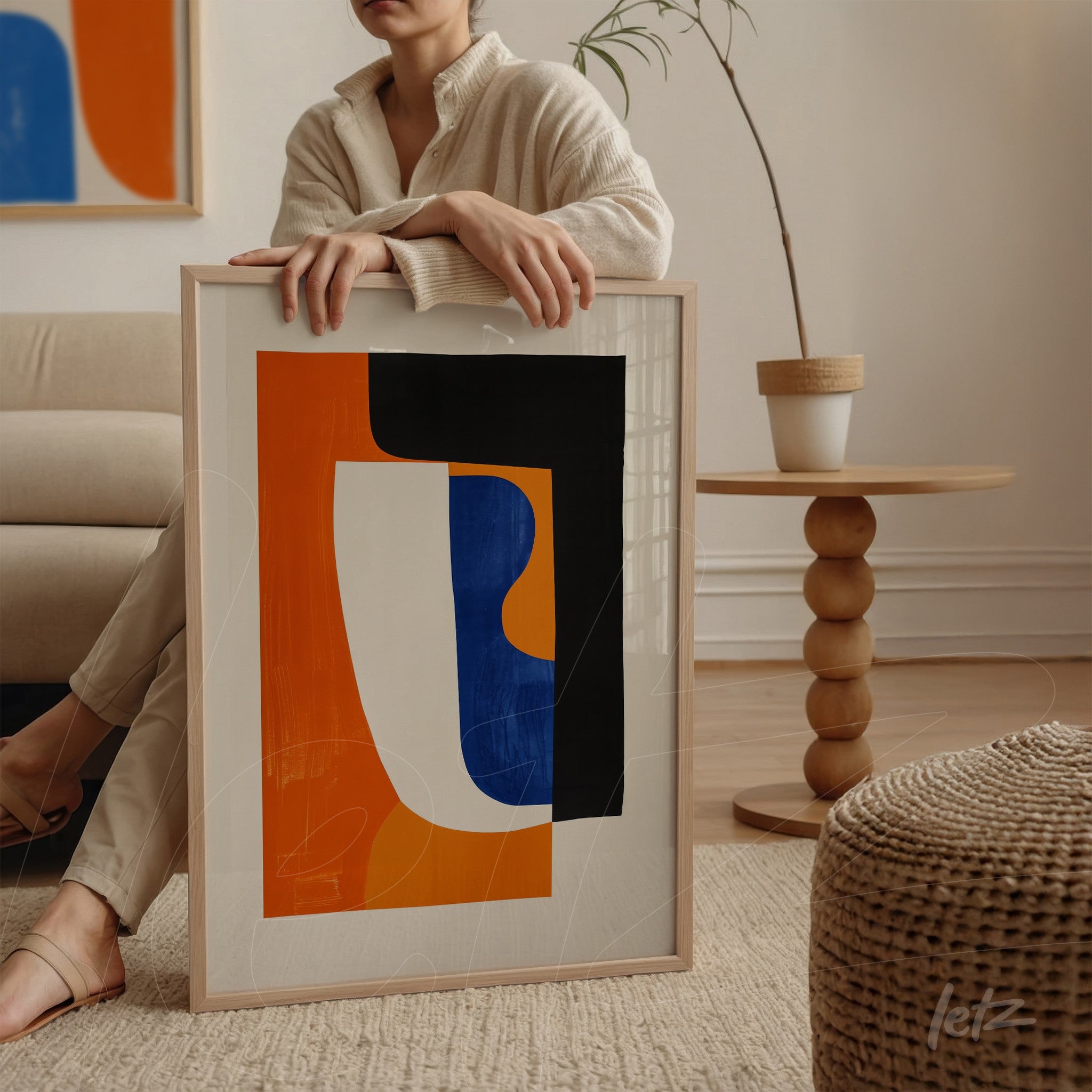 quadro moderno com formas abstratas em tons de laranja, branco e preto, em moldura clara, apoiado ao lado de uma mesa redonda