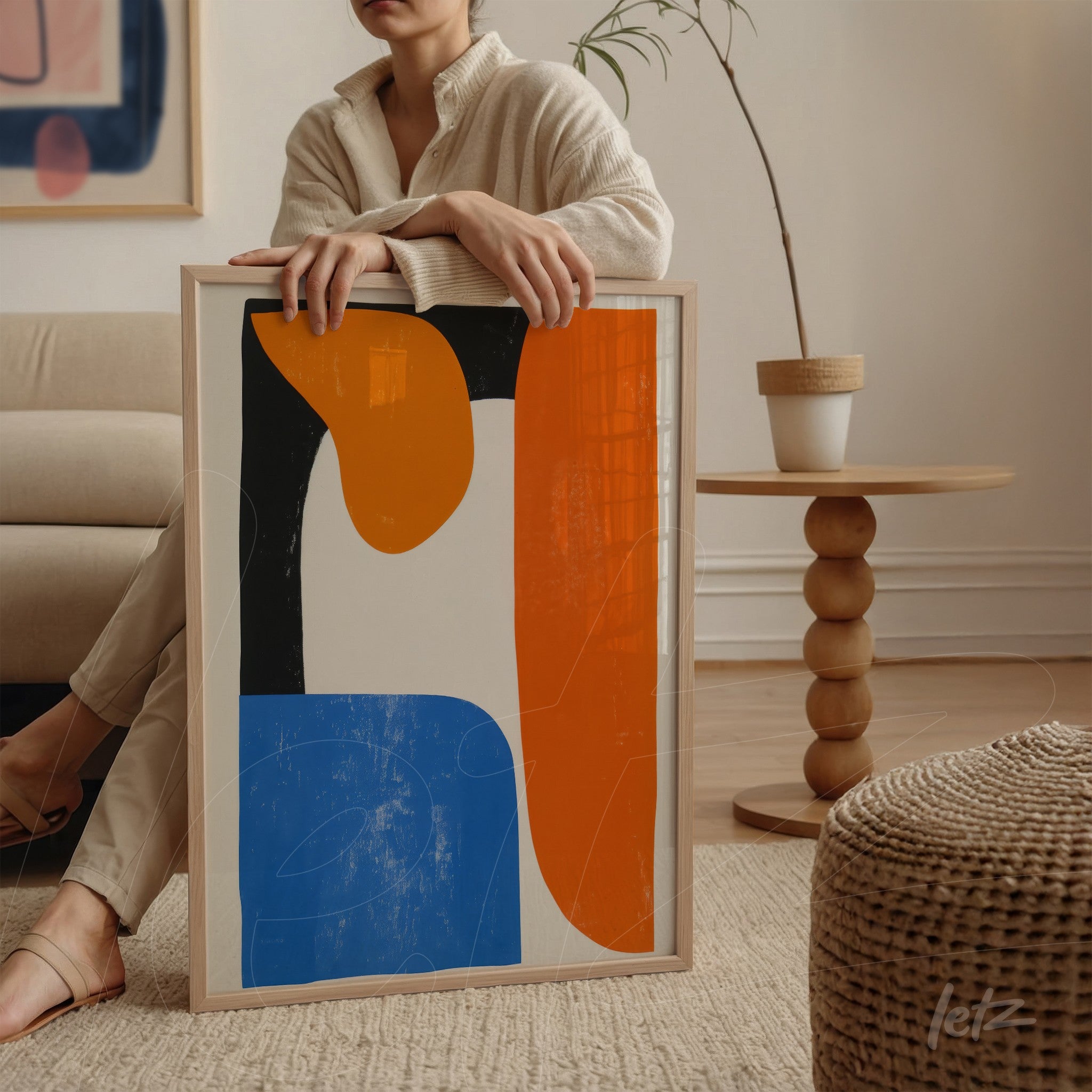 quadro com arte abstrata em tonalidades de azul, laranja e preto, em moldura clara, exibido por uma pessoa sentada ao lado de um sofá