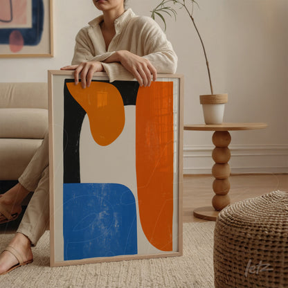 quadro com arte abstrata em tonalidades de azul, laranja e preto, em moldura clara, exibido por uma pessoa sentada ao lado de um sofá