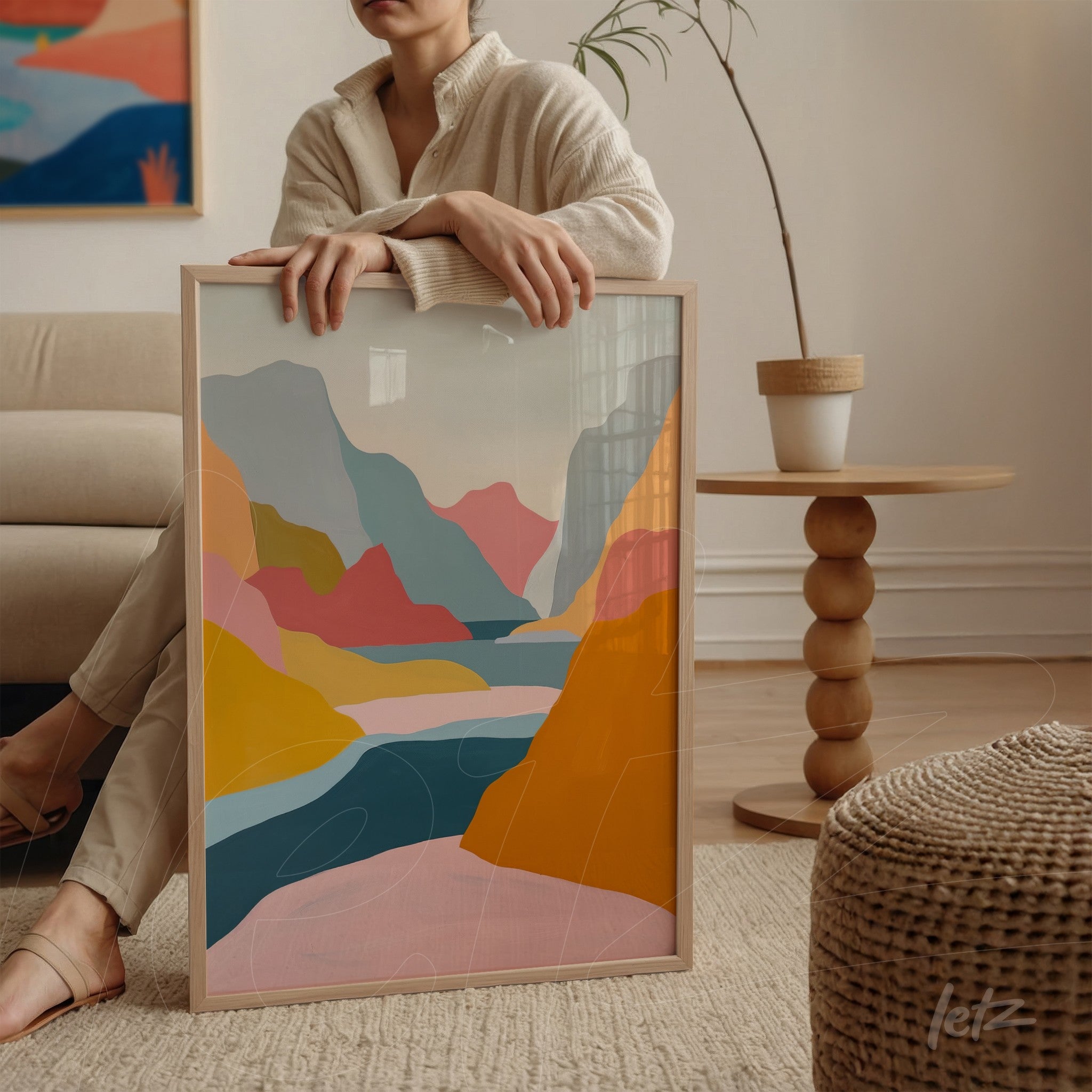 quadro com arte abstrata em tons de azul, rosa e laranja em moldura clara, apoiado em mesa ao lado de uma mulher