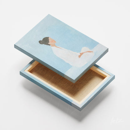 quadro em canvas com figura feminina sentada em meditação, fundo azul suave e moldura clara
